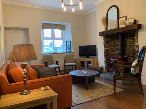 Living room at Ruby Stove Cottage with orange sofas facing the red wood-burning stove in its stone fireplace